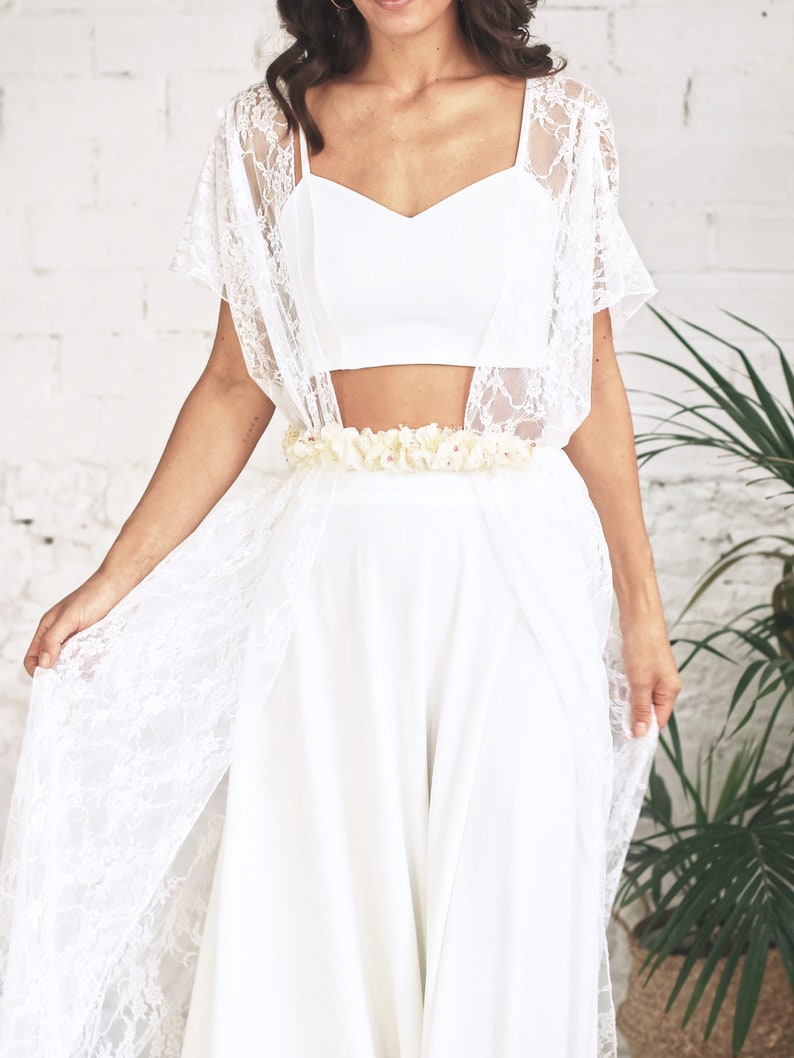 May include: A white lace bridal top with a white crop top underneath and a white flower belt. The top is worn over a white skirt.