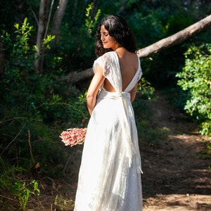 We see a bride in the forest. She is in a natural environment full of light. We see the back of the dress, which can be wrapped in multiple ways. The dress has an ivory base and a lace overlay. The dress has a rustic vibe.