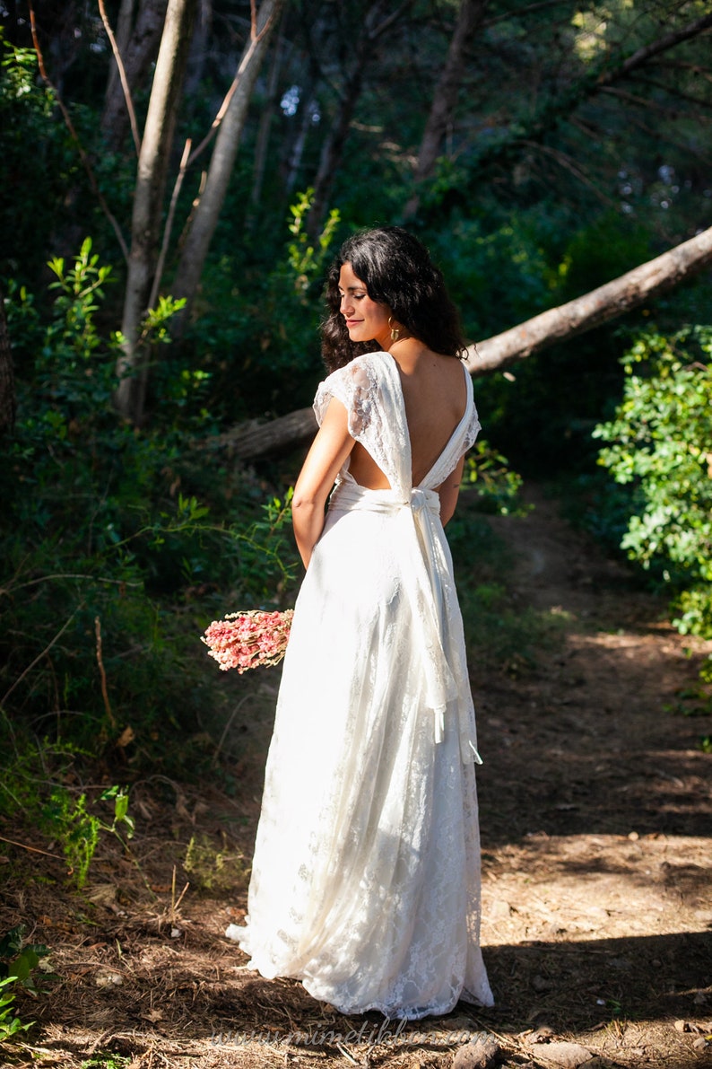 We see a bride in the forest. She is in a natural environment full of light. We see the back of the dress, which can be wrapped in multiple ways. The dress has an ivory base and a lace overlay. The dress has a rustic vibe.