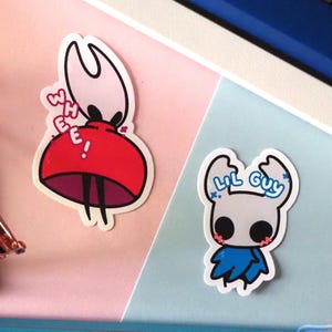 Può includere: Due adesivi con personaggi dei cartoni animati. Uno è rosso e bianco con la scritta "WEE!" e l'altro è bianco e blu con la scritta "LIL GUY". Gli adesivi sono su una superficie rosa e blu con matite e una tastiera sullo sfondo.