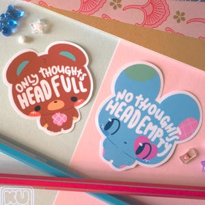 Può includere: Due adesivi fustellati con disegni di animali dei cartoni animati. Uno è un orso marrone con la scritta "ONLY THOUGHTS HEAD FULL". L'altro è un coniglietto blu con la scritta "NO THOUGHTS HEAD EMPTY". Gli adesivi sono su uno sfondo pastello.
