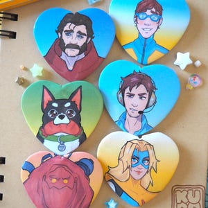 Könnte beinhalten: Sechs herzförmige Knöpfe mit farbenfrohen Illustrationen. Die Designs umfassen Porträts von Personen und einem Hund, jeweils mit einer einzigartigen Farbpalette. Die Knöpfe sind auf einer hellbraunen Oberfläche angeordnet, mit kleinen dekorativen Gegenständen verstreut.