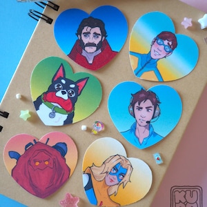 Könnte beinhalten: Eine Sammlung herzförmiger Aufkleber mit Illustrationen verschiedener Charaktere. Die Aufkleber zeigen eine Reihe von Farben, darunter Blau, Rot und Gelb. Die Aufkleber sind auf einem braunen Notizbuch angeordnet, zusammen mit einem Stift und dekorativen Elementen.