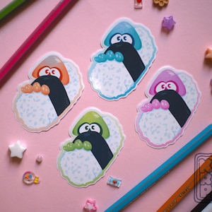 May include: Four colorful stickers featuring cartoon sushi characters with expressive eyes and colorful accents. Each sticker has a different color scheme: orange, blue, pink, and green. The stickers are on a pink background with pencils and decorative elements.
