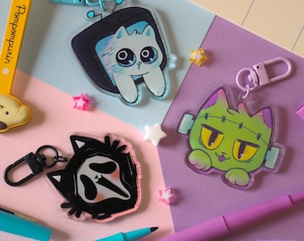 May include: Three acrylic keychains featuring cartoon cats: one white cat inside a television, one green Frankenstein-inspired cat with stitches, and one black cat wearing a ghost mask. Each keychain has a colored lobster clasp attached.