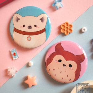 Puede incluir: Dos insignias redondas con diseños de animales de dibujos animados. Una presenta un perro blanco con un collar marrón sobre un fondo azul. La otra muestra un búho marrón sobre un fondo rosa. Elementos decorativos rodean las insignias.