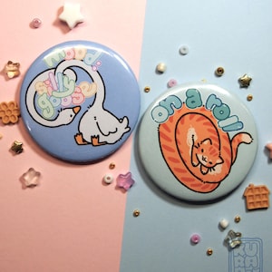 Könnte beinhalten: Zwei runde Ansteckbuttons. Einer ist hellblau mit einer weißen Gänse-Illustration und den Worten "moody goose". Der andere ist hellblau mit einer orangefarbenen Katzen-Illustration und den Worten "on a roll".