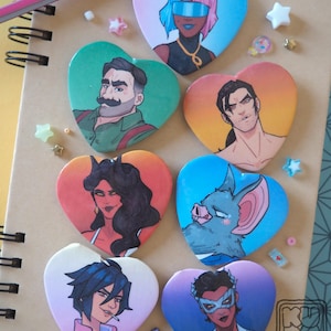 Puede incluir: Colección de botones en forma de corazón con ilustraciones coloridas de personajes. Los diseños varían, incluyendo retratos y figuras de fantasía. El fondo incluye un cuaderno y elementos decorativos.