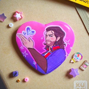 May include: A heart-shaped button with a portrait of a man with long hair and a beard, set against a pink gradient background. The man is wearing a purple jacket and red scarf. The button is surrounded by star-shaped and candy-themed embellishments.