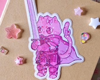 May include: A die-cut sticker featuring a pink cat character wearing dark pink armor and a cape, holding a long, light purple sword. The sticker has a holographic glitter finish with star-shaped sparkles throughout the design.