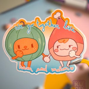 Può includere: Un adesivo colorato con due personaggi dei cartoni animati vestiti da prosciutto e melone, che si tengono per mano. Il testo "we go together like ham and melon" è scritto in un carattere stravagante. Il design è carino e giocoso.
