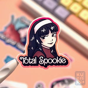 a gradient orange + pink background sticker depicting Tomie from the manga by Junji Ito, smiling,  with the "total spookie" writing below.