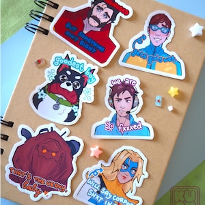 Puede incluir: Una colección de pegatinas coloridas con ilustraciones de dibujos animados y texto. Las pegatinas representan varios personajes y frases, incluyendo "UNDIAGNOSED", "goodest boy" y "gets girls wet". Están dispuestas sobre un cuaderno marrón.