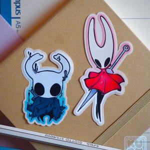 Peut inclure: Deux autocollants en vinyle représentant des personnages du jeu vidéo Hollow Knight. L'un représente un personnage blanc avec une cape bleu foncé. L'autre montre un personnage en cape rouge avec une épée. Les autocollants sont sur un cahier brun.