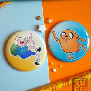 Può includere: Due bottoni rotondi con personaggi di Adventure Time. Un bottone raffigura Finn, l'umano, con camicia e pantaloncini blu, con uno zaino verde. L'altro bottone raffigura Jake, il cane, con corpo arancione e sfondo blu.