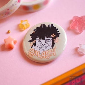 Può includere: Un bottone rotondo bianco sporco con un'illustrazione a cartoni animati di una persona con i capelli neri e la scritta "GULASH" in arancione. Il bottone è circondato da elementi decorativi, tra cui abbellimenti a forma di stella e nastro adesivo colorato.