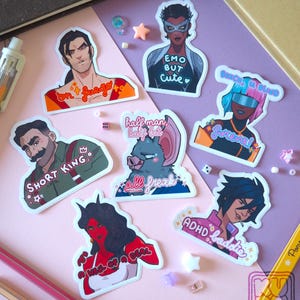 Puede incluir: Una colección de pegatinas coloridas con ilustraciones de personajes estilizados. Las pegatinas incluyen textos como "EMO BUT Cute", "on fuego", "SHORT KING", "hall man half bat", "foreva!" y "ADHD baddie". Están sobre una superficie morada y rosa.