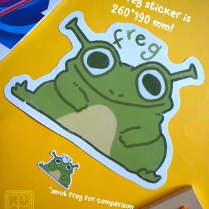 May include: A large green "freg" sticker, 260mm x 190mm, with a cartoon frog design, is displayed on a yellow surface. A smaller version of the sticker is also present for size comparison. The word "freg" is written above the frog.