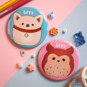 May include: Two circular pin badges featuring cartoon animal illustrations. One is light blue with a dog, the other is pink with an owl. Both have the word "bffs" and are surrounded by small decorative items.