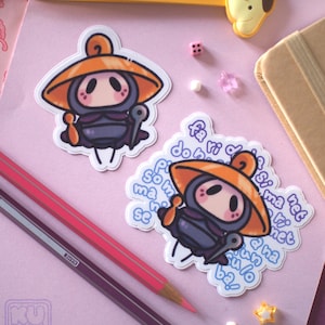 Puede incluir: Dos pegatinas con un personaje de dibujos animados con sombrero naranja y atuendo morado. Las pegatinas están sobre una superficie rosa con lápices, un accesorio Pompompurin amarillo y otros pequeños objetos decorativos.