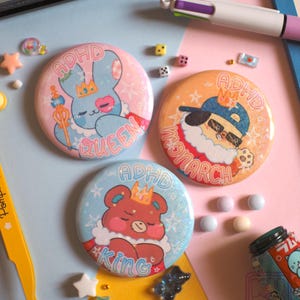Peut inclure: Trois badges ronds avec des illustrations de dessins animés. L'un représente un lapin bleu avec une couronne et le texte "ADHD QUEEN". Un autre montre un chien avec des lunettes de soleil et "ADHD MONARCH". Le troisième présente un ours avec "ADHD KING".