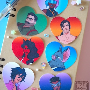 May include: A collection of heart-shaped stickers featuring various illustrated characters. The stickers showcase a diverse range of styles and colors, with characters in different poses and outfits. The stickers are on a notebook.