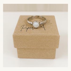 Puede incluir: Un anillo plateado con una gema blanca y redonda se muestra sobre una pequeña caja de cartón marrón. La caja tiene la palabra "Piper" escrita en cursiva. El anillo presenta un diseño minimalista.