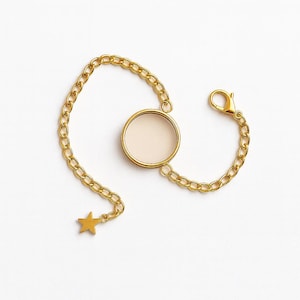 Puede incluir: Pulsera de cadena dorada con un colgante circular y un charm de estrella. El colgante tiene un centro color crema. La pulsera presenta un cierre de mosquetón y está sobre un fondo blanco.