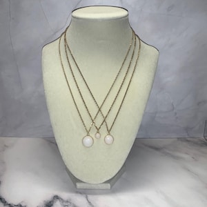 May include: Three gold-toned necklaces with white circular pendants displayed on a cream-colored bust. The necklaces vary in length, with the pendants hanging at different levels. The bust is set against a marble-patterned background.