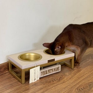May include: A white and gold elevated pet food and water bowl set with two bowls. The bowls are made of stainless steel and have a gold finish. The stand is made of wood and has a white finish. The stand has a personalized name tag that says "YOUR PET'S NAME". A brown cat is eating from one of the bowls.