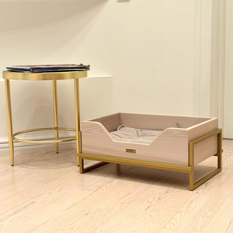 May include: A light wood dog bed with a gold metal frame. The bed has a soft, beige cushion inside. The bed is sitting on a light wood floor.