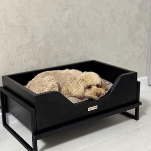 May include: A black wooden dog bed with a raised frame and a beige cushion. A small brown dog is lying in the bed. The bed has a small wooden plaque with the word "BARLEY" on it.