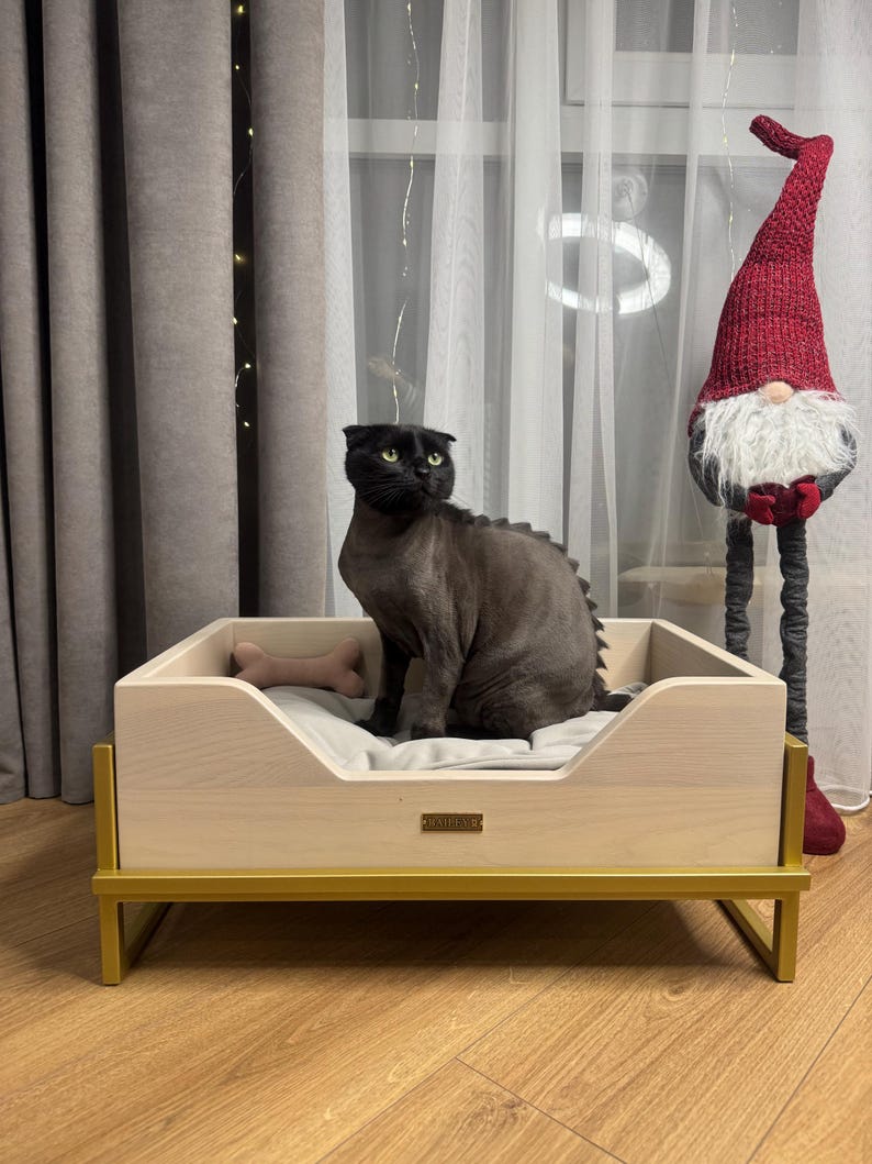 May include: A black cat sits in a light wood dog bed with a gold metal frame. The bed has a gray cushion and a bone-shaped toy. The bed is on a wood floor.