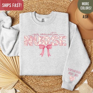 May include: A gray sweatshirt with the text "air evacuation NURSE" in pink floral letters with a pink bow. The text "SAMANTHA BSN, RN" is on the sleeve with a pink heart.