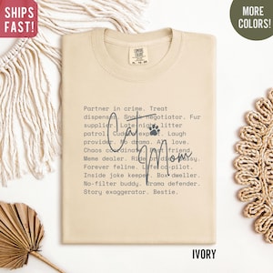 May include: Ivory-colored t-shirt with the text "Cat Mom" and a paw print. The shirt lists various cat-related roles such as "Partner in crime," "Snack negotiator," and "Forever feline."
