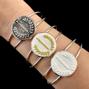 May include: Three silver bangle bracelets with a circular design featuring a sword and laurel wreath. The sword and wreath are in different colors on each bracelet: silver, gold, and white.