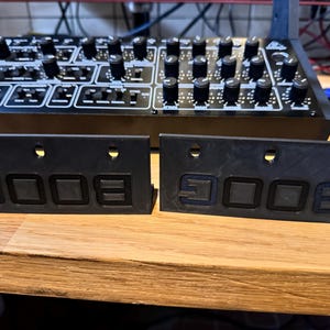 Könnte beinhalten: Zwei schwarze Paneele mit dem Wort "BOOG" in fetter Schrift. Sie befinden sich vor einem schwarzen Synthesizer mit zahlreichen Knöpfen und Bedienelementen. Der Hintergrund ist eine Holzoberfläche.