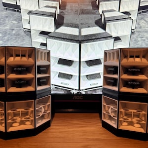 Puede incluir: Dos grandes gabinetes de altavoces negros y blancos con múltiples altavoces en cada gabinete. Los altavoces están dispuestos en un patrón hexagonal.