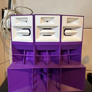 May include: Three purple and white speaker cabinets stacked on top of each other. The speakers have a square shape and are made of wood. The speakers are connected to each other by a metal frame.