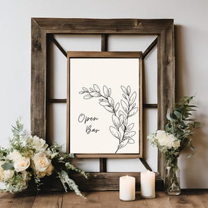 Puede incluir: Un cartel imprimible con una ilustración en blanco y negro de una rama frondosa y el texto "Open Bar" en una fuente sencilla. El cartel está enmarcado en un marco de madera rústico y se muestra en una mesa con flores blancas y velas.