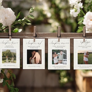 May include: Four cards with wedding photos and text are clipped to a wooden board. The cards display phrases such as "Happily Surprised," "Engaged Life," and "The Wedding Chapter." White flowers and greenery surround the cards, creating a decorative display.