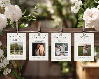 May include: Four rectangular cards with photos and text are clipped to a wooden board. The cards feature wedding-related photos and phrases like "Happily Surprised," "Engaged Life," and "The Wedding Chapter." White flowers and greenery frame the scene.