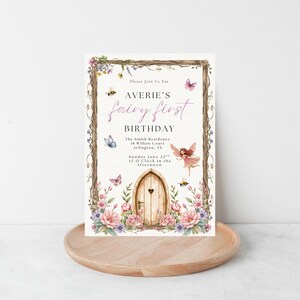 Puede incluir: Una invitaci&oacute;n de cumplea&ntilde;os con tem&aacute;tica de hadas. La invitaci&oacute;n presenta una puerta de madera con un coraz&oacute;n, rodeada de flores, mariposas y abejas. El texto dice "Averie's fairy first birthday". La invitaci&oacute;n est&aacute; sobre una bandeja de madera.