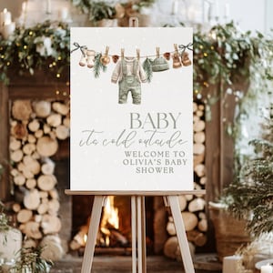 May include: A baby shower sign on a wooden easel. The sign features a watercolour illustration of baby clothes hanging on a washing line. Text on the sign reads "BABY it's cold outside" and "WELCOME TO OLIVIA'S BABY SHOWER". The background is a soft, speckled white.
