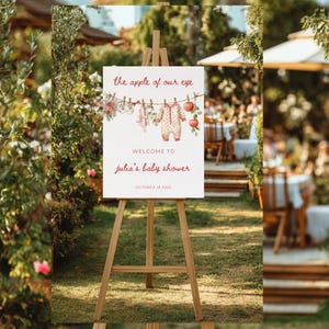 May include: A wooden easel displays a baby shower welcome sign. The sign reads "the apple of our eye" above a clothesline with baby clothes and apples. Below, it says "Welcome to Julia's baby shower" and the date.