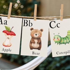 Puede incluir: Tres tarjetas del alfabeto se muestran, cada una sujeta por una pinza de madera. Las tarjetas presentan ilustraciones de una manzana, un oso y una oruga, con las letras y palabras correspondientes: A, Apple; B, Bear; C, Caterpillar. El fondo está borroso.