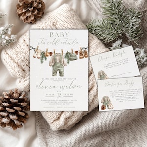 May include: Baby shower invitation set with a winter theme. The invitation features watercolour illustrations of baby clothes hanging on a washing line. Includes a diaper raffle card and a book request card. The invitation is surrounded by pine cones and winter greenery.