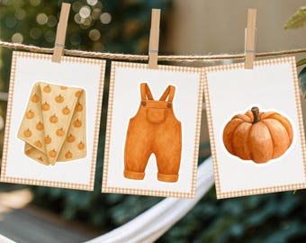 May include: Three watercolor illustrations of autumn-themed items are displayed on a clothesline. The images include a folded towel with pumpkin designs, orange overalls, and a pumpkin. Each image has a white background with a light orange gingham border.