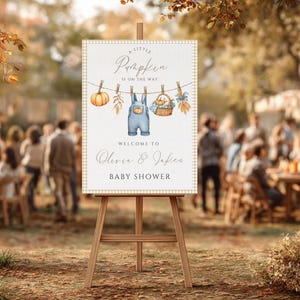 May include: A baby shower sign on a wooden easel. The sign features a pumpkin, overalls, and a basket illustration. Text reads "A Little Pumpkin Is On The Way" and "Welcome to Olivia & Jake's Baby Shower."
