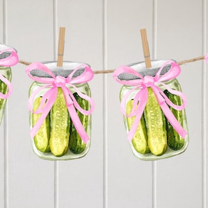 May include: Watercolor illustration of glass jars filled with pickles, each adorned with a pink ribbon and a wooden clothespin. The jars hang from a rope against a white background.